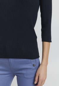 Granatowy sweter z prążkowanym splotem i rękawami do trzech czwartych, w parze z jasnoniebieskimi spodniami z dwoma dekoracyjnymi guzikami po bokach.