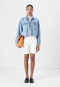 Chaqueta de denim con mangas largas, dos bolsillos en el pecho, usada sobre una blusa beige. Pantalones cortos blancos y mocasines negros. Sosteniendo un bolso naranja.