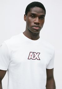 Mladi crni muškarac nosi bijelu majicu s podebljanim "AX" logom u crvenoj i crnoj boji na prsima, neutralna svijetla pozadina.