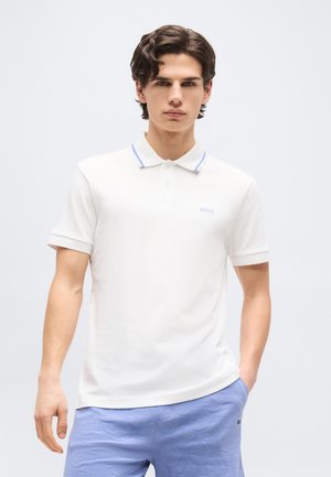 Νεαρός άνδρας με κοντά σγουρά μαλλιά που φοράει λευκό πόλο μπλουζάκι με μπλε ροζάκια και ανοιχτό μπλε υφασμάτινο σορτς, στέκεται μπροστά από μια απλή, μονόχρωμη επιφάνεια.