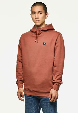 Brauner Hoodie mit Kordelzug, gerippten Bündchen und einem kleinen schwarzen Logo-Patch auf der Brust, kombiniert mit blauen Jeans. Weicher, strukturierter Stoff.