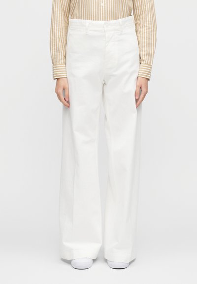 Hvite bukser med vide ben laget av bomull med en glatt tekstur og høyt liv. Brukes med en stripete langermet skjorte og hvite sko.