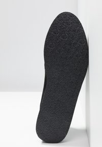 Semelle de chaussure en caoutchouc noir avec un motif texturé antidérapant, photographiée de face sur un fond blanc avec des coins.