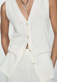 Biały, dziergany, bezrękawnik z głębokim dekoltem w kształcie litery V, zapinany na guziki, z delikatnymi wzorami tekstury; cechuje się luźnym krojem i bocznymi rozcięciami.