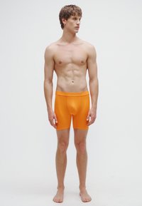 Oranžové boxerky z elastickej látky, s priliehavým strihom a mäkkým pásom. Model pózuje na boso.