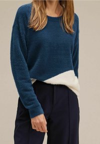 Kvinna klädd i en blå och vit fluffig tröja med långa ärmar och mörka byxor, stående mot en enfärgad beige bakgrund.