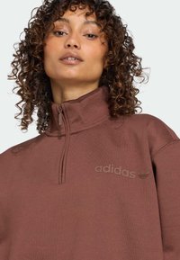 Barna, cipzáros kapucnis pulóver magas gallérral, minimalista dizájnnal és a mellkasán lévő, azonos árnyalatú, dombornyomott Adidas logóval.