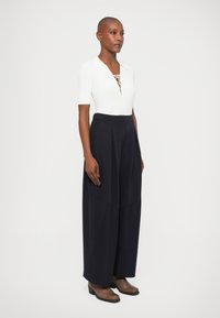 Witte geribde top met korte mouwen en een vetersluiting bij de hals, gecombineerd met losse, marineblauwe high-waisted broek. Bruine enkellaarsjes maken de outfit af.