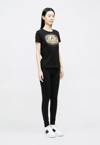 Fekete póló "EA7 Emporio Armani" logóval, fekete leggingsszel és fekete részletekkel díszített fehér tornacipőkkel párosítva. Simaságú anyag, testhezálló fazon.