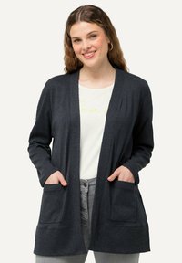 Mörkgrå cardigan med ribbad struktur, öppen frontdesign och två framfickor. Bärs över en ljus t-shirt och ljusa jeans.