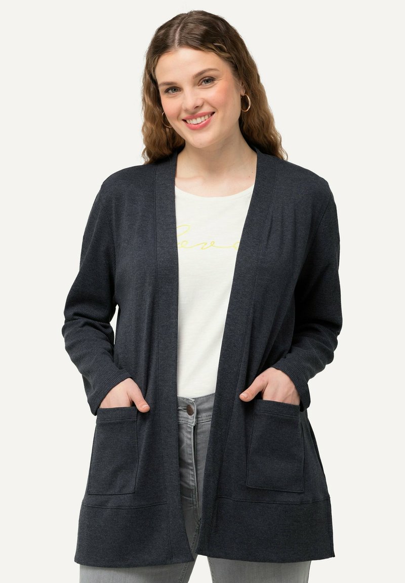 Mörkgrå cardigan med ribbad struktur, öppen frontdesign och två framfickor. Bärs över en ljus t-shirt och ljusa jeans.