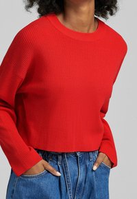 Czerwony prążkowany sweter z okrągłym dekoltem i długimi rękawami, zestawiony ze swobodnymi niebieskimi dżinsami. Tkanina wygląda na miękką i teksturowaną.