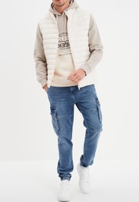 Chaleco ligero y acolchado sobre una sudadera con capucha beige, combinado con pantalones cargo azules y zapatillas blancas. Conjunto casual en capas.