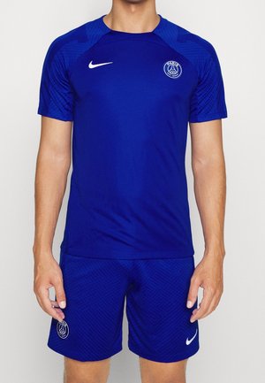 Uomo che indossa la divisa da calcio blu del Paris Saint-Germain con loghi Nike su maglia e pantaloncini, in piedi davanti a uno sfondo uniforme.