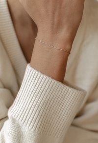 Brazalete delgado y delicado en metal rosado, con un diseño retorcido. Lleva puesto en una muñeca, complementado con un suéter de punto texturizado claro.