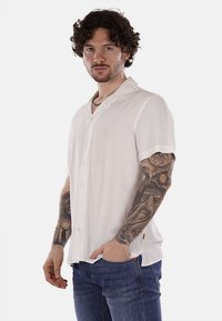 Camicia bianca a maniche corte con bottoni, realizzata in tessuto leggero, caratterizzata da un colletto a punte e morbidi dettagli nei bottoni. Indossata con pantaloni jeans blu.
