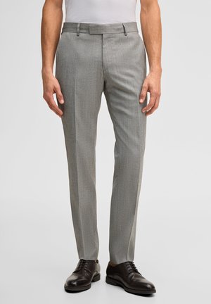 Uomo con pantaloni eleganti grigi aderenti e scarpe stringate in pelle nera, in piedi contro uno sfondo semplice.