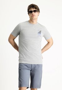 Camiseta de algodón gris con mangas cortas y un diseño gráfico azul en el bolsillo, combinada con pantalones cortos de color azul claro. Hombre con gafas de sol negras.