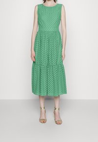 Vestido verde sin mangas con un diseño de ojales texturizados, falda de capas y escote redondo. Combinado con tacones dorados de tiras.