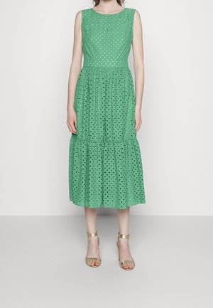 Robe verte sans manches avec un design texturé en trou d'œil, jupe à volants et encolure ronde. Associée à des talons dorés à lanières.