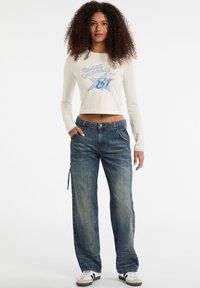 Junge Frau mit lockigem Haar, die ein weißes Langarm-Crop-Top mit einem Sternen-Graphic und lockeren blauen Jeans mit weißen Sneakers trägt.