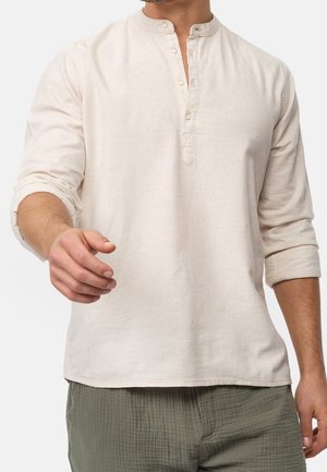 Mann trägt ein beigefarbenes Langarm-Henley-Shirt mit hochgekrempelten Ärmeln und olivgrüne, strukturierte Hosen, Hand nach vorne gestreckt.