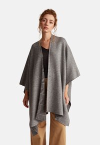 Šedý vlněný pončo s volným, zavěšeným designem, s houndstooth vzorem podél okrajů. Měkká textura a otevřený přední díl.