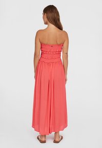 Abito maxi senza spalline di color corallo con corpino arricciato, gonna fluida e dettagli a volant, realizzato in tessuto leggero. Sandali piatti visibili sotto.