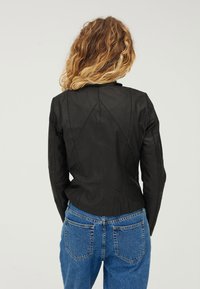 Kvinna med lockigt blont hår klädd i en tight svart läderjacka och blå jeans, vänd bort från kameran mot en ljus, enkel bakgrund.