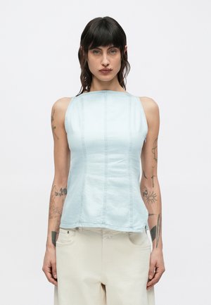 Person med mørkt hår iført lyseblå ermeløs topp og lyse beige bukser, med tatoveringer på begge armer, stående foran en enkel bakgrunn.