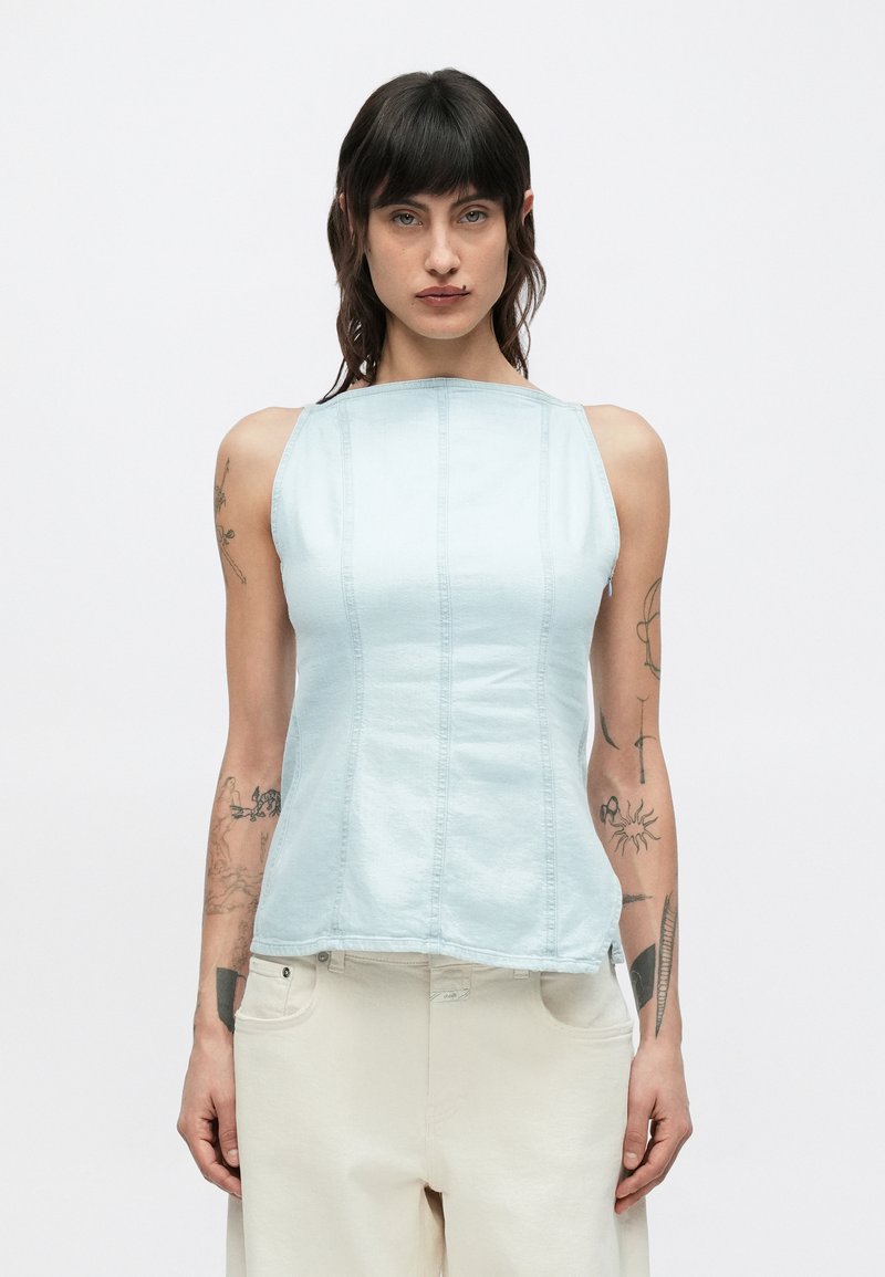 Άτομο με σκουρόχρωμα μαλλιά που φοράει αμάνικο γαλάζιο τοπ και ανοιχτό μπεζ παντελόνι, με τατουάζ και στα δύο χέρια, όρθιο μπροστά σε απλό φόντο.