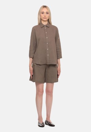 Donna con capelli biondi lisci indossa una camicia marrone con bottoni e maniche a tre quarti, abbinata a pantaloncini coordinati e scarpe basse scure.