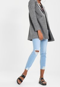 Šedý texturovaný blazer s klopovým límcem, nošený přes černý květinový top. Světle modré džínsy s opotřebeným vzhledem a černé sandály dokončují outfit.
