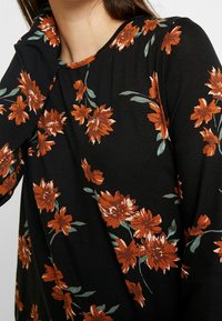 Camisa negra de manga larga con estampado floral naranja y hojas verdes, llevada por una persona que se toca el cuello, recortada por encima de la boca.