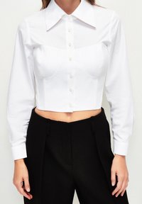 Camicetta bianca corta con un design strutturato, dotata di colletto a punta, chiusura con bottoni e maniche lunghe. Abbinata a pantaloni neri a vita alta.