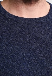 Gros plan d'une personne portant un pull à col rond texturé bleu foncé, montrant le cou et le haut de la poitrine.