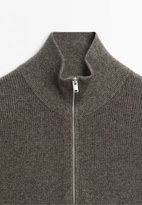 Närbild av en brun ribbad stickad tröja med hög krage och en delvis uppdragen silverfärgad dragkedja.