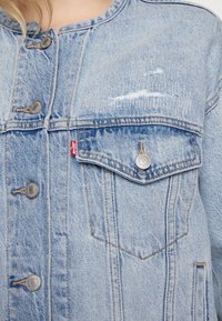 Lichtblauwe spijkerjack met zilveren knopen, borstzak en versleten details. Rechte snit met zichtbare stiksels en naadaccenten.