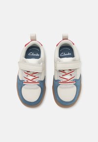 Hvit og blå lærsko med røde kontraster, med borrelås, snøring og en beige gummisåle. Clarks-merke synlig.
