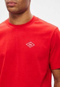 Άνδρας με φωτεινό κόκκινο μπλουζάκι με λαιμόκοψη και μικρό λευκό διαμαντένιο λογότυπο στο αριστερό στήθος.