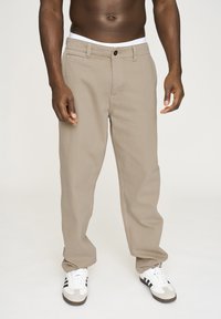 Beige bomuldsbukser med lige ben, der har knaplukning, bæltestropper og forlommer. Bæres med hvide og sorte sneakers.