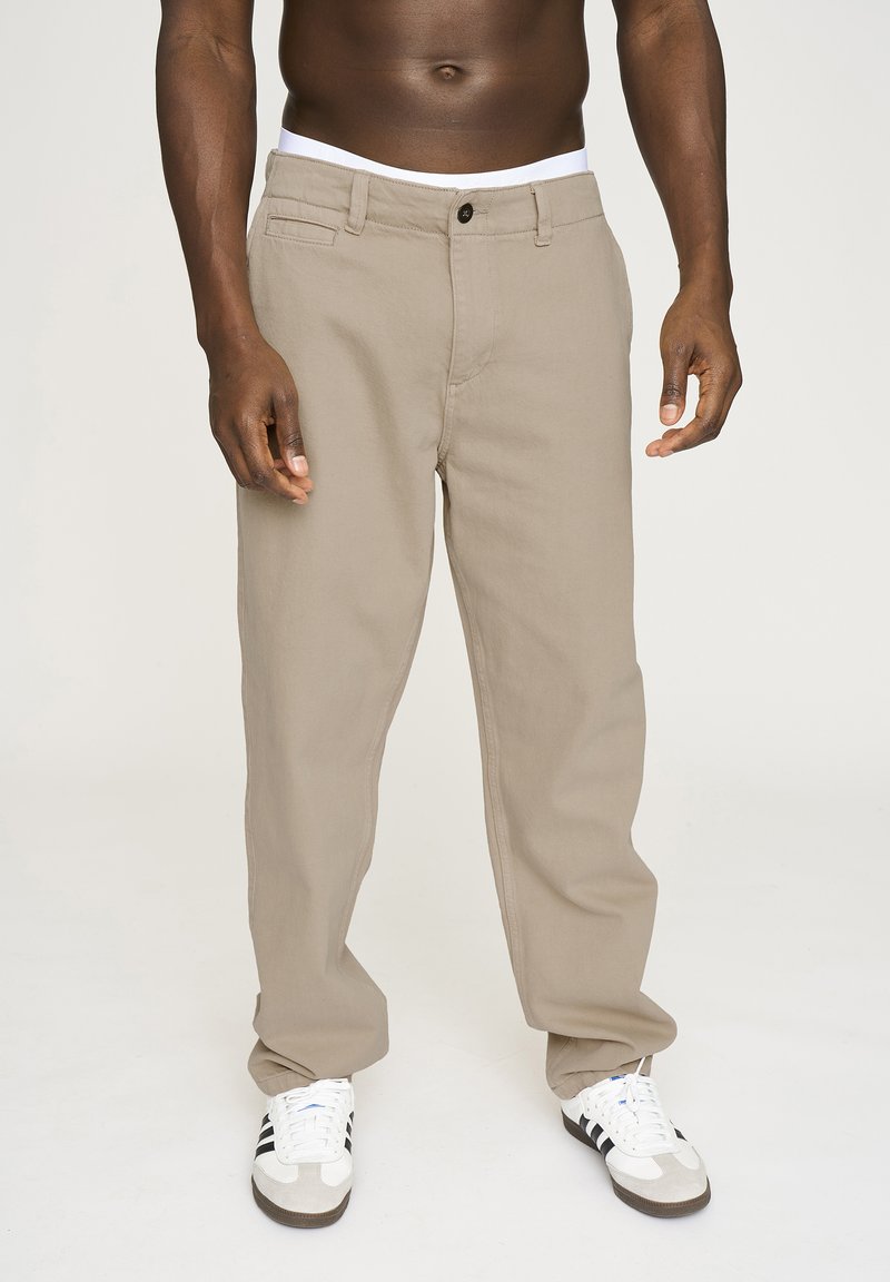 Beige bomuldsbukser med lige ben, der har knaplukning, bæltestropper og forlommer. Bæres med hvide og sorte sneakers.