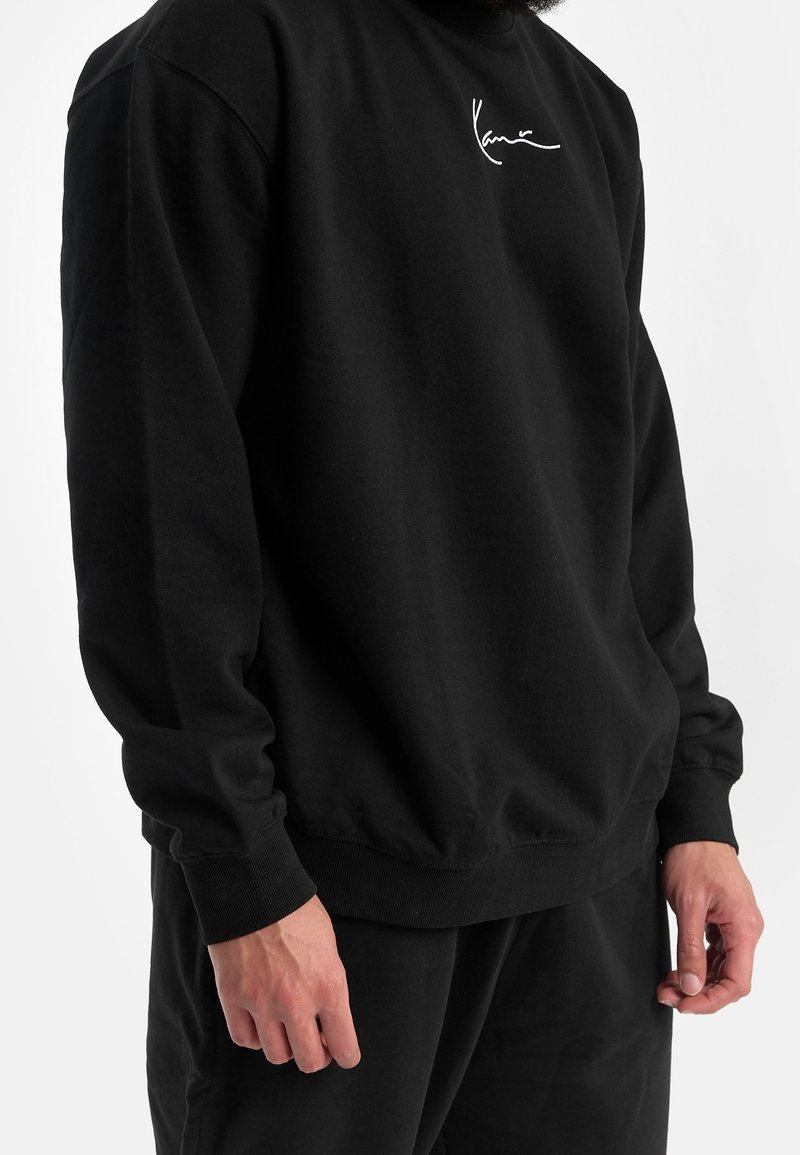 Homme portant un sweat-shirt noir à manches longues et un pantalon, logo blanc sur le haut gauche de la poitrine, debout les mains détendues le long du corps.