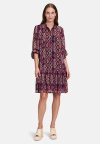 Short-sleeve, knee-length dress in pink, purple, and cream, featuring a geometric pattern, button detailing, and a ruffled hemline.