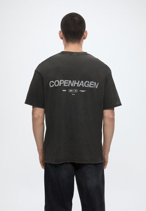 Mężczyzna noszący czarną koszulkę z napisem „COPENHAGEN” i małym logo na środku górnej części pleców, stojący na tle jednolitego jasnego tła.