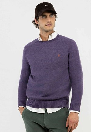 Hombre con suéter de cuello redondo morado sobre una camisa blanca, pantalones verde oscuro y gorra de pana negra, mirando hacia un lado.