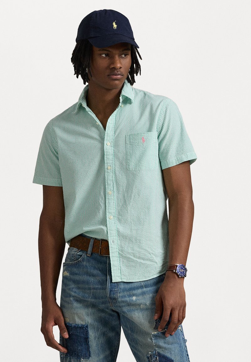 Camicia a maniche corte a righe verdi e bianche con taschino a sinistra e logo discreto, abbinata a jeans blu strappati e un cappellino blu navy.