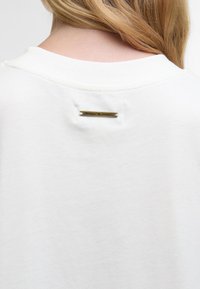 Pessoa loira a usar uma camisa branca com uma pequena etiqueta metálica perto do pescoço com a inscrição "Barbour ♥ FARM RIO."