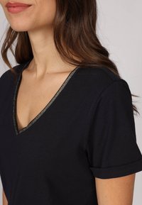 T-shirt noir à col en V en tissu doux, avec un accent doré autour de l'encolure et des manches courtes retroussées pour un ajustement décontracté.