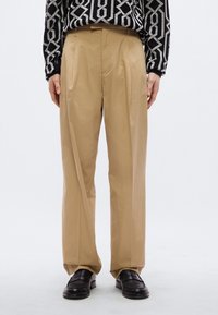 Μπεζ πλισέ παντελόνια με λείο υφή και άνετη εφαρμογή. Συνδυασμένα με μαύρα loafers και μαύρο τοπ με σχέδιο.